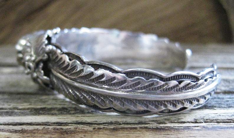 May include: A silver bracelet with a feather design. The bracelet is made of sterling silver and has a detailed feather pattern.