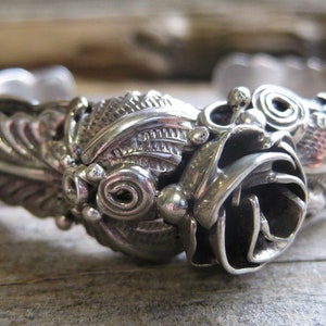 May include: A silver bracelet with a detailed rose design. The rose is made of intricate swirls and has a textured surface.