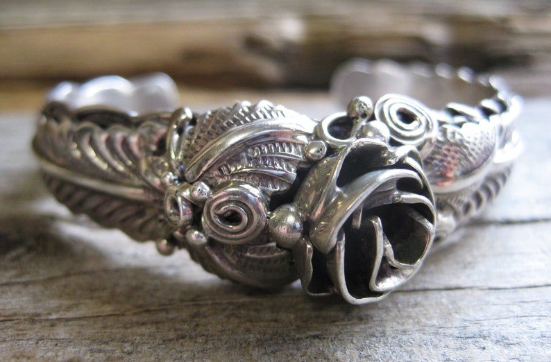 May include: A silver bracelet with a detailed rose design. The rose is made of intricate swirls and has a textured surface.