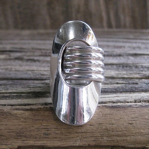 Könnte beinhalten: Ein silberfarbener Ring mit ovaler Form und einem spiralförmigen Design in der Mitte. Der Ring hat eine polierte Oberfläche, die das Licht reflektiert. Das Design ist modern und geometrisch.