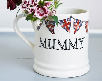 May include: White ceramic mug with a handle, decorated with a string of red, white, and blue bunting flags and the word "MUMMY" in black lettering. A bouquet of pink and red carnations is in the mug.
