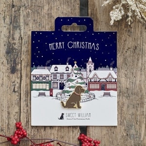 Puede incluir: Un pin de esmalte con un perro sentado frente a un árbol de Navidad en una escena de pueblo nevado. El pin dice "Merry Christmas" y "Sweet William" en la tarjeta de respaldo.