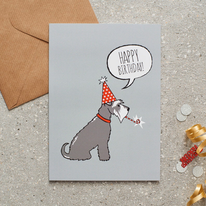 May include: A gray birthday card featuring a cartoon dog wearing a party hat and holding a party favor. The card has the words "HAPPY BIRTHDAY!" in a speech bubble. An envelope and party decorations are in the background.