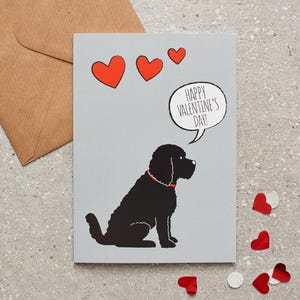 Puede incluir: Tarjeta de San Valentín con una ilustración de un perro negro con un collar rojo y tres corazones rojos. La tarjeta tiene un bocadillo que dice "Happy Valentine's Day!" y viene con un sobre marrón.