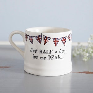 May include: White ceramic mug with a handle, featuring a string of bunting with Union Jack flags and the text "Just HALF a Cup for me DEAR..."
