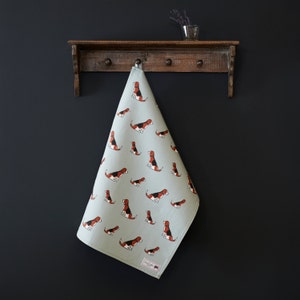 May include: A light blue tea towel with a repeating pattern of brown and black beagle dogs. The tea towel is hanging on a wooden rack with hooks.