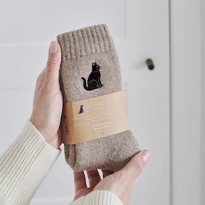Könnte beinhalten: Beige Wollsocken mit einer schwarzen Katze mit rotem Halsband aufgestickt. Die Socken werden von einer Person gehalten. Ein braunes Papierband um die Socken zeigt den Schriftzug "Sweet William" und eine Hunde-Silhouette.
