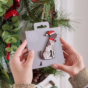 Pode incluir: Um íman de Natal com um cão com um gorro de Pai Natal e uma campainha, apresentado num cartão cinzento. O cartão diz "Merry Christmas" e "Sweet William".