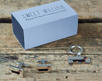 May include: A set of dachshund-themed gifts in a silver box. The set includes a keychain, cufflinks, and a small box. The dachshund is black with tan markings.