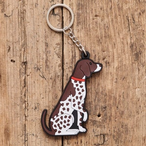 Peut inclure: Un porte-clés en forme de chien tacheté brun et blanc avec un collier rouge. Le chien est assis, la tête tournée vers la droite. Le porte-clés est en caoutchouc et possède un anneau en métal argenté.