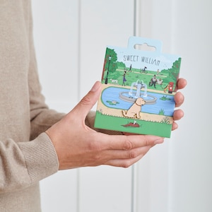 Puede incluir: Una tarjeta con una ilustración de dibujos animados de un perro sentado junto a una fuente en un parque. La tarjeta es verde y azul con el texto "Sweet William" en la parte superior.