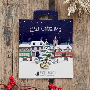 Könnte beinhalten: Eine goldfarbene Emaille-Anstecknadel mit einem Hund, der vor einem Weihnachtsbaum in einer verschneiten Dorfszene steht. Die Anstecknadel trägt die Aufschrift "Frohe Weihnachten" und "Sweet William Home of the Mischievous Mutts".