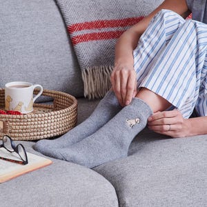 Può includere: Un paio di calzini grigi con un piccolo cane giallo ricamato su una calza. I calzini sono indossati da una persona seduta su un divano grigio.