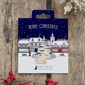 Puede incluir: Pin de esmalte dorado con un perro sentado frente a una escena de pueblo nevado con un árbol de Navidad. El pin tiene un fondo blanco con las palabras "Merry Christmas" y "Sweet William" impresas en él.