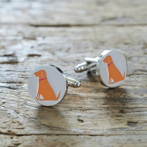 Könnte beinhalten: Ein Paar silberne Manschettenknöpfe mit weißem Hintergrund und einer Illustration eines orangefarbenen Hundes. Der Hund trägt ein rotes Halsband.