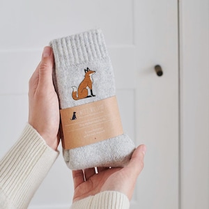 Könnte beinhalten: Hellgraue Socken mit einer Fuchsgrafik und dem Text "SWEET WILLIAM". Die Socken sind mit einem braunen Papierband umwickelt. Der Fuchs ist braun und weiß.