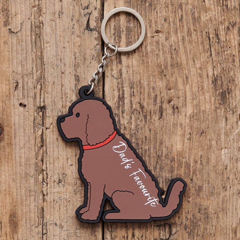 以下が含まれることがあります： 座っている犬の形をした茶色のゴム製のキーホルダーで、側面に「Dad's Favourite」という文字が書かれています。