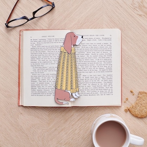 May include: A brown and white bloodhound dog wearing a yellow and brown sweater. The dog is looking to the right. The dog is a bookmark and is placed in an open book.