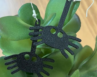 May include: A pair of black earrings with a unique design featuring a stylized eye with multiple rays emanating from it. The earrings are made of a textured material and have silver hooks.