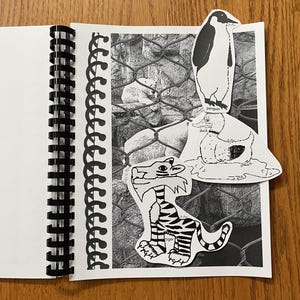 May include: A black and white notebook with a spiral binding. The notebook has a black and white photo of a polar bear in a cage. There are three cut-out images of animals on the page: a penguin, a duck, and a tiger. The penguin is standing on top of the duck, and the duck is standing on top of the tiger. The tiger is labeled "tiger" and the duck is labeled "duck". The penguin is labeled "penguin".