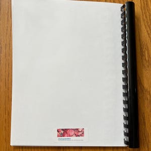 May include: A white notebook with a black plastic binding. The notebook has a sticker on the front that says "Original Artwork" and features a pink and red abstract design.
