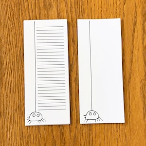 May include: Two white rectangular notepads with a black spider illustration at the bottom. One notepad has horizontal lines, the other is blank. A black line extends from the spider to the top of each notepad. The notepads are on a wooden surface.