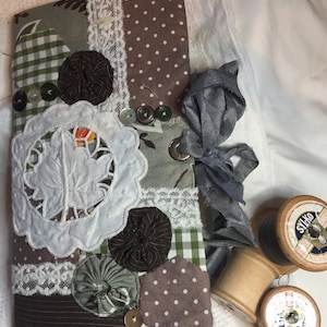 Puede incluir: Una bolsa de tela de patchwork marrón y verde con ribete de encaje, botones y una flor de tela blanca. La bolsa está cerrada con una cinta gris y está sobre una superficie blanca junto a dos carretes de hilo de madera.