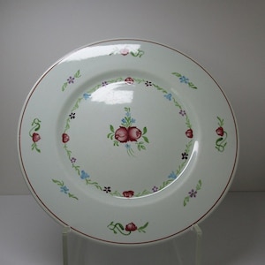 May include: White ceramic dinner plate with a brown rim and a floral design in shades of pink, blue, green, and purple. The design features a central cluster of pink roses with green leaves and smaller flowers around the edge.
