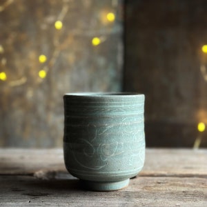 May include: A light blue ceramic teacup with a delicate white design. The cup is sitting on a rustic wooden surface.