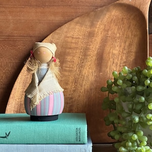 May include: A wooden doll with a pink and gray striped dress, a white crocheted shawl, and a white hat with red pom-poms. The doll is sitting on a stack of books with the text "HOUGHTON MIFFLIN CO." visible.