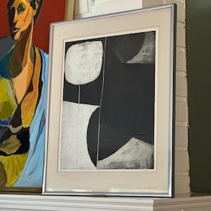 May include: Framed abstract art print with a silver metal frame. The artwork features a black and white geometric design with rounded shapes and lines. The print is displayed on a white surface, with a colorful painting visible in the background.