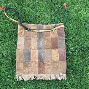 May include: A patchwork suede shoulder bag in shades of brown, tan, and gray, with a fringed bottom. The bag has a rectangular shape and is set against a backdrop of green grass. The straps are black and green.
