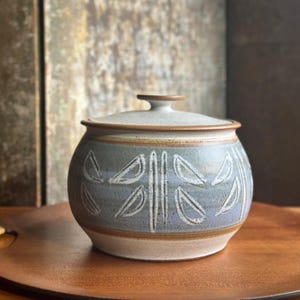 May include: A ceramic jar with a lid, featuring a light blue and white color scheme. The jar has a rounded shape with a lid and is decorated with leaf-like patterns. The jar is sitting on a wooden tray.