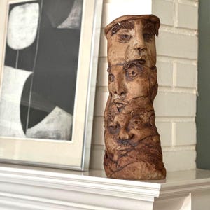 May include: A tall, textured ceramic sculpture featuring stacked faces with detailed features. The faces are in varying sizes and expressions, with a natural, earthy brown color. The artwork is displayed on a white surface.
