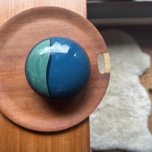 May include: A round wooden serving tray with cut-out handles holds a blue and green ceramic sphere. The sphere is divided by a black line. The tray is on a wooden surface.