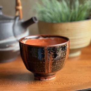 May include: A handmade ceramic tea bowl with a dark brown exterior and a reddish-brown interior. The bowl has a slightly irregular shape and a glossy finish. A black teapot and a decorative plant are in the background.