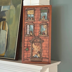 May include: A rectangular, wooden wall hanging depicting a brick building facade with windows and a door. The windows show figures, and the door has the number 303. The artwork is framed in a dark brown wood.