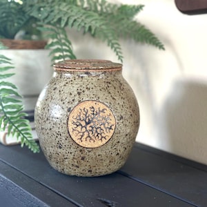 May include: A speckled brown ceramic jar with a lid, featuring a stylized tree design in a circle on the front.
