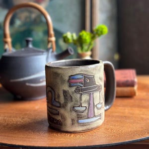 May include: A ceramic mug with a Libra zodiac design. The mug is tan with a dark brown handle and rim. The Libra symbol is depicted in a stylized illustration with red, blue, and purple accents. A teapot and books are in the background.