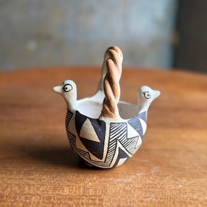 May include: A small, handcrafted ceramic basket with a twisted handle. The basket features two bird-like figures on either side, with black and white geometric patterns adorning the body. The handle and the birds' beaks are a light brown color.