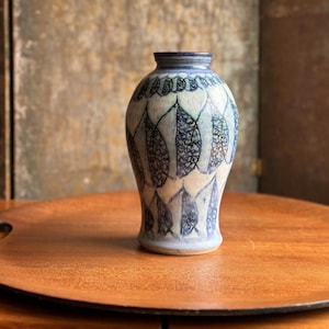 May include: A ceramic vase with a tapered neck and a rounded body, featuring a light cream base color decorated with repeating dark blue leaf-shaped patterns. The surface displays a textured, hand-painted finish with intricate blue line details.