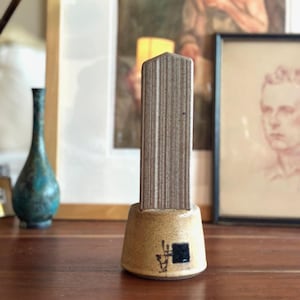 May include: A ceramic obelisk-shaped sculpture with a textured, vertical-lined surface in shades of brown and tan. The base is a lighter tan color with a black square and abstract markings. The sculpture is displayed on a wooden surface.
