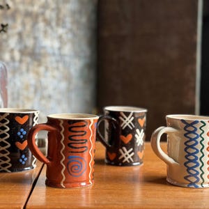 May include: Four ceramic mugs with unique hand-painted designs. The mugs feature various patterns, including hearts, zigzags, and swirls, in colors like orange, blue, and white. Each mug has a handle and sits on a wooden surface.