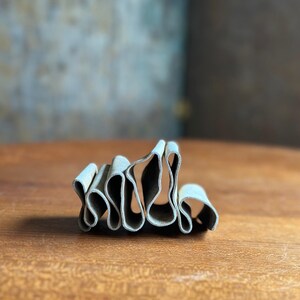 May include: A small, abstract sculpture with a wavy, layered design. The piece is made of a light gray material and sits on a wooden surface. The sculpture's form creates a play of light and shadow.