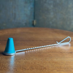 May include: A bar spoon with a teal cone-shaped end and a twisted silver handle. The spoon rests on a wooden surface, with the handle extending to a looped end. The teal end is a solid color.