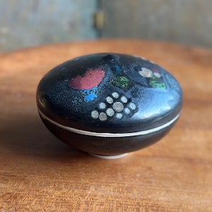 May include: A small, black, oval-shaped ceramic trinket box with a white band around the middle. The lid is decorated with colorful abstract designs in red, blue, green, and white. The box sits on a wooden surface.