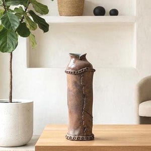 May include: A tall, cylindrical ceramic vase with a mottled brown glaze. The vase features a textured, raised band around the neck and base, with a vertical seam-like detail running down the side. The rim has a subtle, pinched, organic shape.