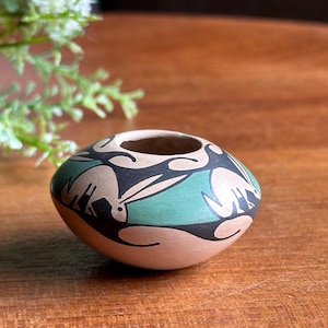 May include: A small, round, terracotta-colored ceramic pot featuring a matte finish. The exterior is decorated with a repeating pattern of stylized black rabbits set against a muted teal green background, with smooth, curved lines encircling the vessel.