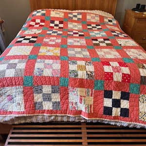 May include: A handmade quilt with a red background and a patchwork design. The quilt features squares of various colors and patterns, including white, black, and teal. The quilt is on a wooden bed frame.