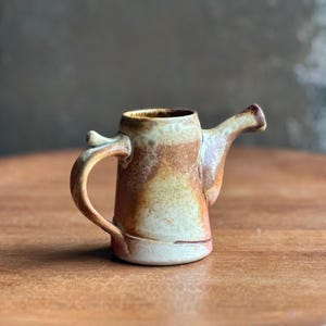 May include: A small, handmade ceramic pitcher with a handle and spout. The pitcher features a rustic design with a blend of brown, beige, and white colors. The handle curves gracefully, and the spout extends outward. The pitcher is likely used for pouring liquids.